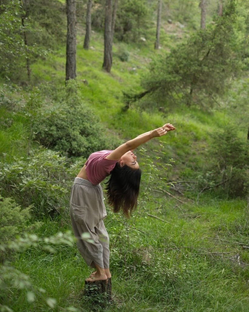 A woman practicing yoga outdoors on a tree stump in a lush green forest. She is performing a backbend stretch, embracing nature and wellness.