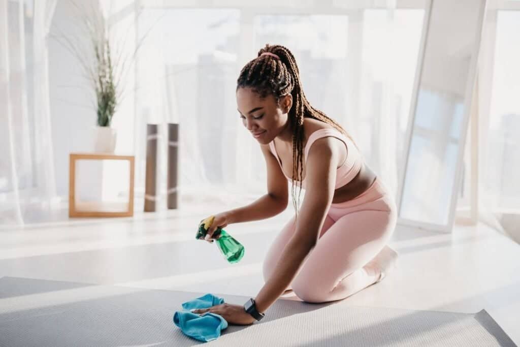 Vibrant young woman cleaning Yoga mat with spray and cloth in sunny home environment, practicing cleanliness and mindfulness for holistic wellness.