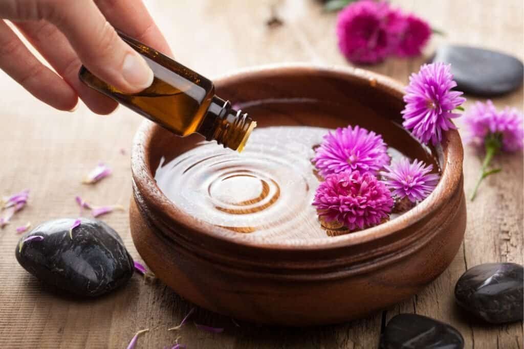 Aromatic essential ayurvedic hair oil being added to a bowl of water surrounded by pink flowers and smooth black stones.