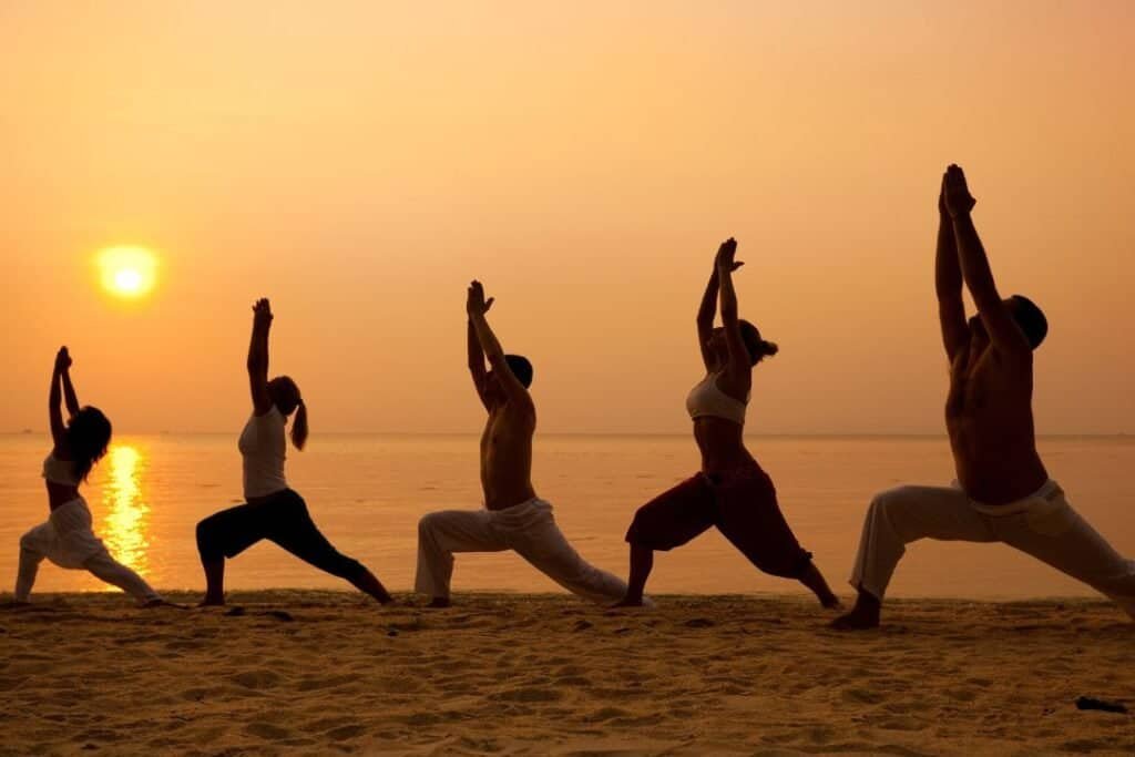 Sunset yoga session on the beach with practitioners performing Warrior pose in silhouette.