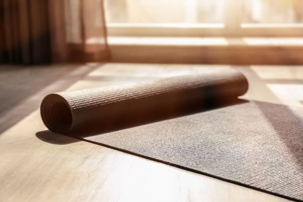 Soft yoga mat rolled out on wooden floor with sunlight streaming through window.