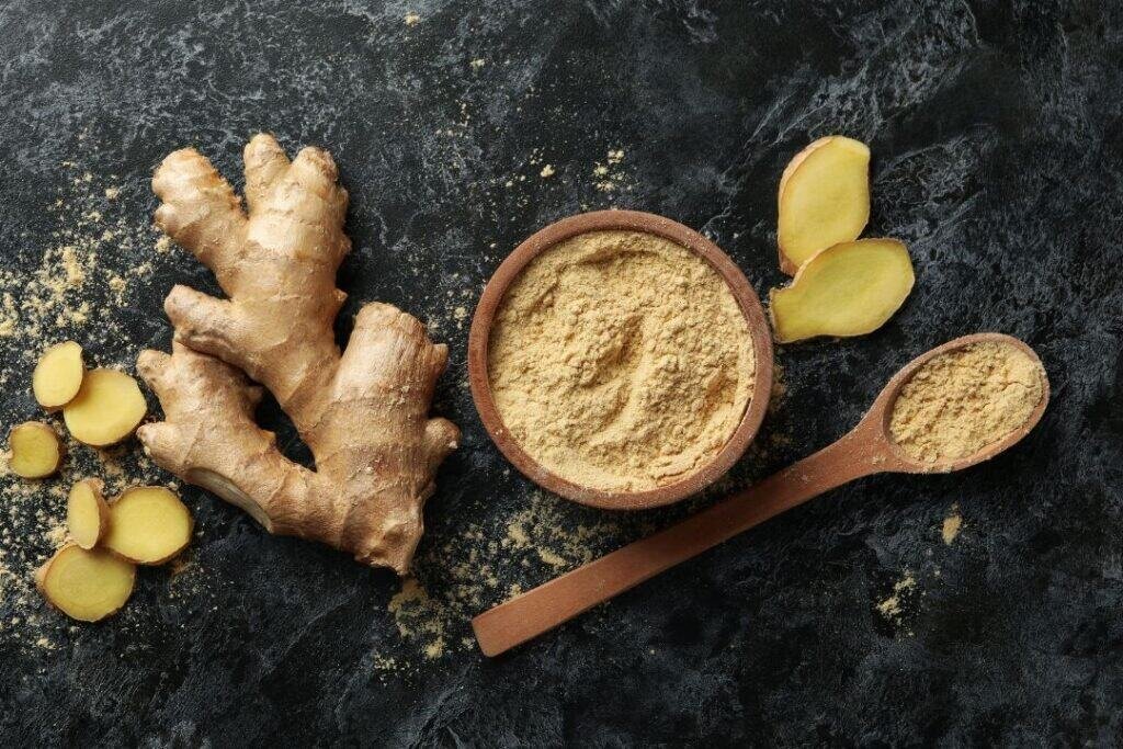 Fresh ginger root, ginger powder in a bowl, and sliced ginger on a dark textured surface, illustrating natural ingredients used in Ayurveda and herbal remedies.