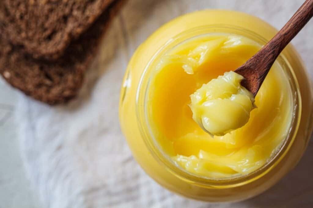 Creamy yellow Ayurvedic organic ghee in a glass jar with a wooden spoon, on a rustic surface with bread slices in the background.